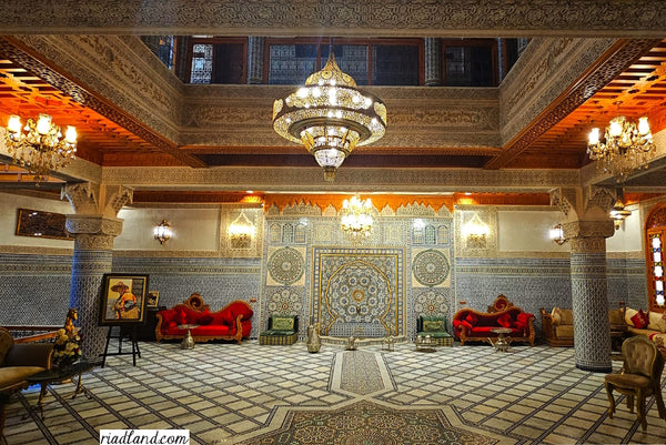 The interior of a luxurious Riad is adorned with colorful zellige tiles and a large brass lamp as the centerpiece.