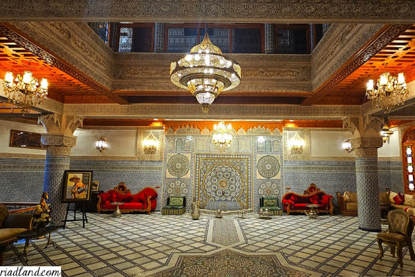 The interior of a luxurious Riad is adorned with colorful zellige tiles and a large brass lamp as the centerpiece