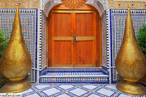 Entrance of a riad with a large wooden door flanked by two traditional standing lamps, featuring handcrafted tiles adorning the floor and wall