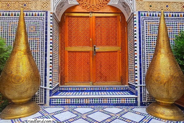 Entrance of a riad with a large wooden door flanked by two traditional standing lamps, featuring handcrafted tiles adorning the floor and wall